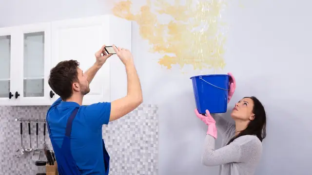 Homeowner photographing a water-stained wall while a bucket catches a leak